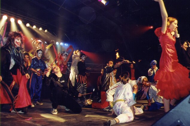 Une douzaine de jeunes aux costumes flamboyants et aux maquillages élaborés dans différentes positions souvent acrobatiques. Au centre, un homme manipule un baton enflammé au deux extrémités. Une douzaine de jeunes aux costumes flamboyants et aux maquillages élaborés dans différentes positions souvent acrobatiques. Au centre, un homme manipule un baton enflammé au deux extrémités.