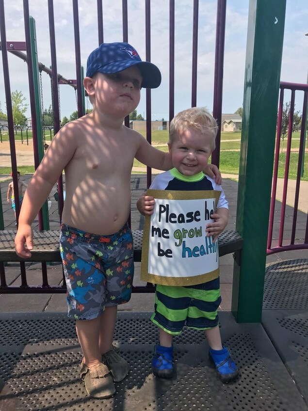 Luke Germs prend la pose à côté d'un garçon né une semaine après lui. Il lui arrive à l'épaule. Il tient une affiche où il est écrit « svp, aidez-moi à grandir en santé ». Ils se trouvent au parc. Luke Germs prend la pose à côté d'un garçon né une semaine après lui. Il lui arrive à l'épaule. Il tient une affiche où il est écrit « svp, aidez-moi à grandir en santé ». Ils se trouvent au parc.