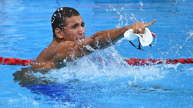 Un nageur célèbre sa victoire dans la piscine et montre du diogt son entraîneur dans les gradins.