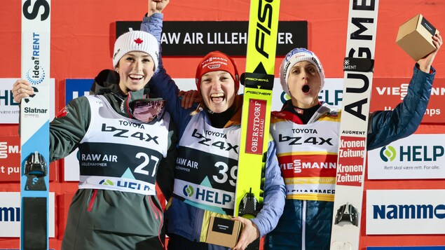 Alexandria Loutitt, à gauche, avec un manteau vert-de-gris et un bandeau blanc avec une feuille d'érable rouge bien centrée, célèbre sa deuxième place, skis blancs à la main, aux côtés de l'Allemande Katharina Althaus et l'Autrichienne Eva Pinkelnig.  