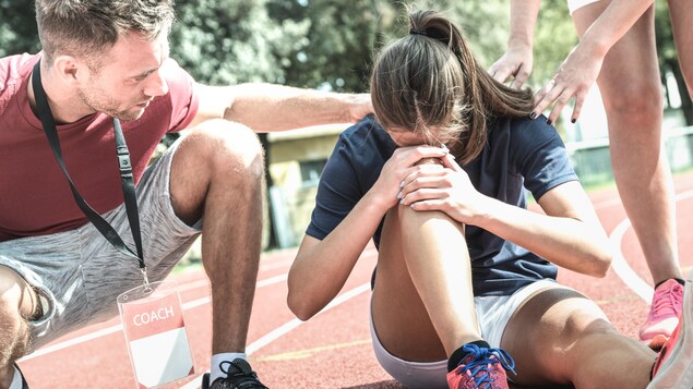 L'adolescente, assise au sol, se tient le genou. Son entraîneur la réconforte.