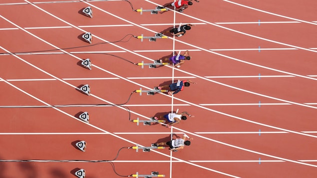 Vue aérienne de six coureurs qui sortent des blocs de départ d'un sprint sur une piste d'athlétisme.