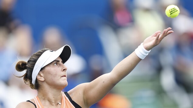 Une joueuse de tennis, vêtue d'une camisole orange, lance la balle pour servir. 