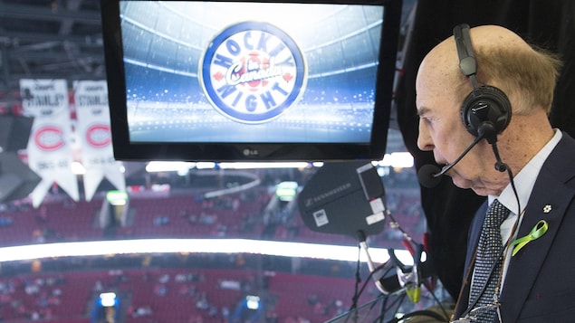 Un homme âgé sur la galerie de presse du Centre Bell, regarde la glace en bas. Il porte des écouteurs et un micro. Il est écrit Hockey Night in Canada sur un écran.