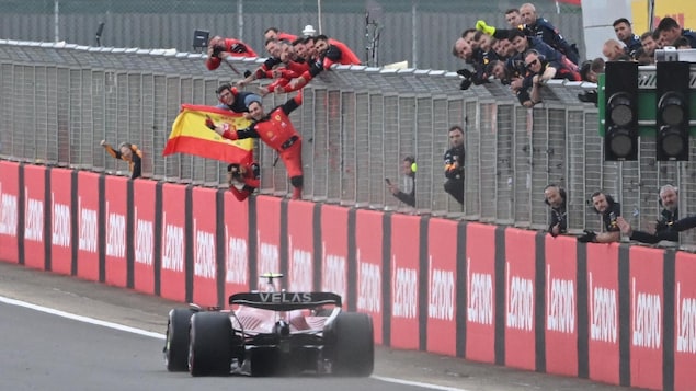 Une monoplace F1, de dos, passe la ligne d'arrivée, des mécaniciens saluent le pilote, un drapeau espagnol flotte.