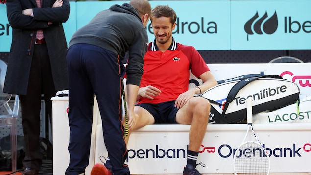 Un joueur de tennis assis en bordure du terrain grimace pendant qu'un  soigneur lui parle.