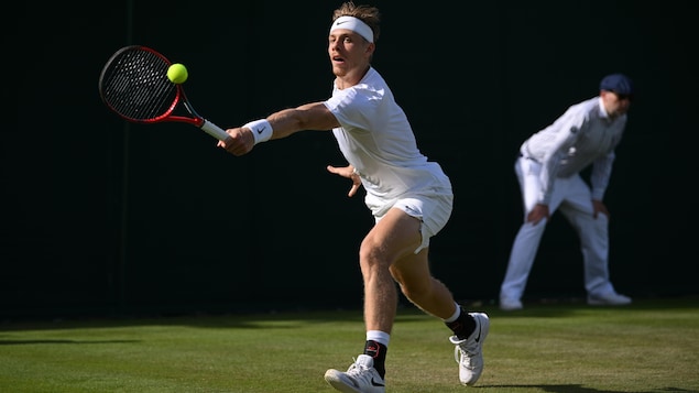 Un joueur de tennis, tout de blanc vêtu, s'étire pour frapper une balle du revers avec sa raquette rouge sur un terrain de gazon. 