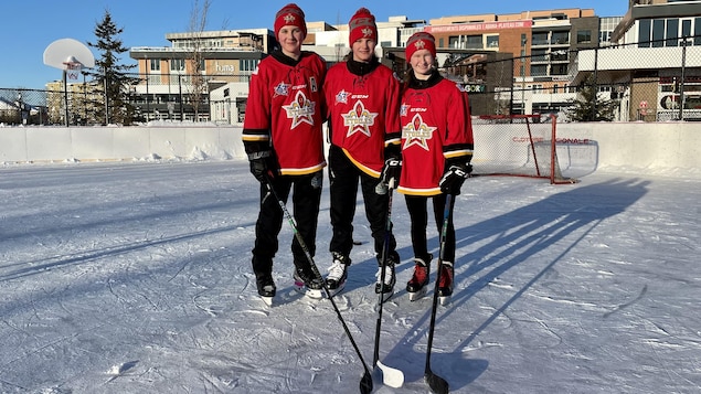 Trois jeunes femmes prennent une photo avec leur bâton de hockey sur une patinoire extérieure.