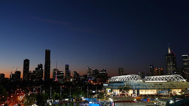 Vue générale du centre-ville de Melbourne le soir, avec un stade éclairé à l'avant-plan.