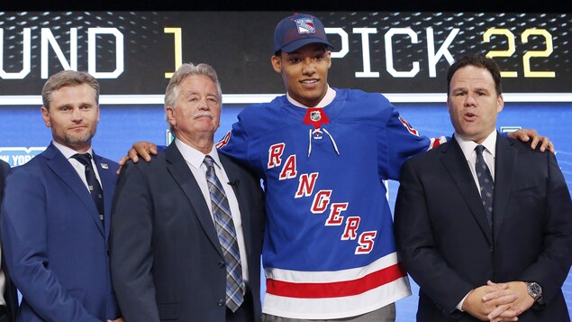 Un jeune joueur de hockey pose pour la photo, vêtu du chandail et de la casquette des Rangers, avec des officiels de sa nouvelle équipe, après avoir été repêché.