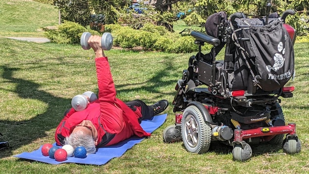 Il est couché au sol avec des haltères, à côté de son fauteuil motorisé.