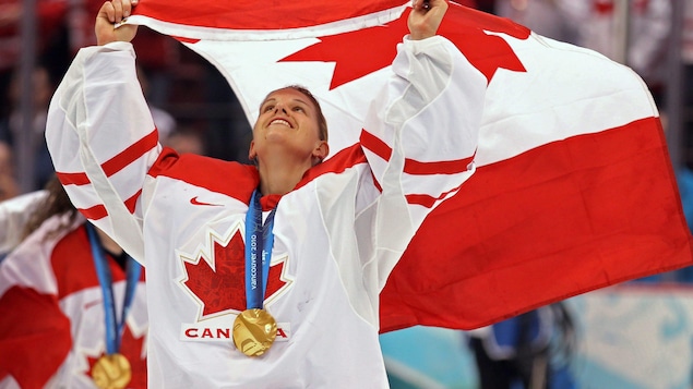 La gardienne de but fait flotter le drapeau canadien au-dessus de sa tête. Elle a une médaille d'or autour du cou.