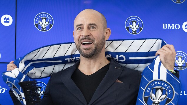 Un homme en veston sourit en tenant une écharpe du CF Montréal.