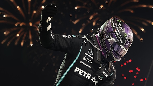Lewis Hamilton, casqué, en combinaison noire, lève le bras droit, il y a des feux d'artifice derrière lui.