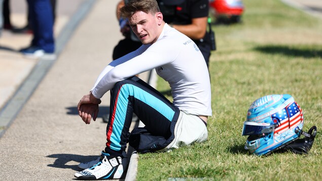 Un pilote de course est assis dans l'herbe et regarde sur sa gauche.