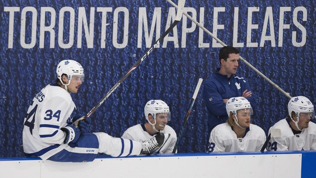 Un joueur de hockey est assis sur la bande alors que ses coéquipiers assis au banc suivent l'action des yeux.