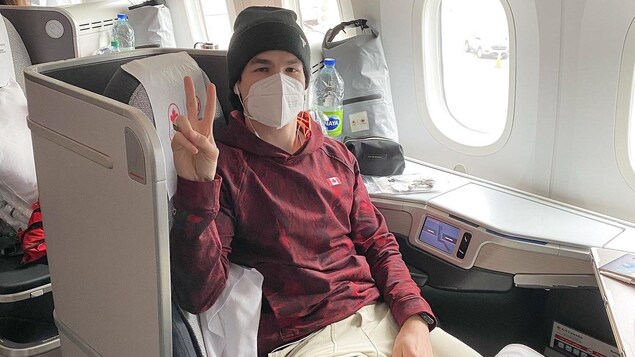 Un homme avec un chandail rouge et un masque blanc fait un signe de victoire assis dans un avion