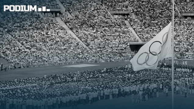 Une photo en noir et blanc d'un grand rassemblement dans un stade, avec à droite le drapeau olympique en berne