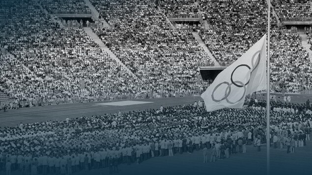 Une photo en noir et blanc d'un grand rassemblement dans un stade, avec à droite le drapeau olympique en berne