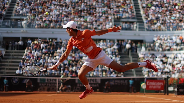 Le joueur de tennis serbe, avec un polo orange et un short et une casquette blanche, saute pour frapper la balle du revers sur la terre battue du terrain principal de Rome. 