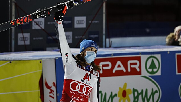 Portant le masque, elle lève son bras droit avec un ski dans la main.