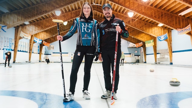 Deux joueurs de curling - un homme et une femme - sourient sur la glace, leur balais à la main.