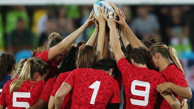 Les joueuses de rugby de l'équipe canadienne se rassemblent au centre du terrain et soulèvent le ballon. 