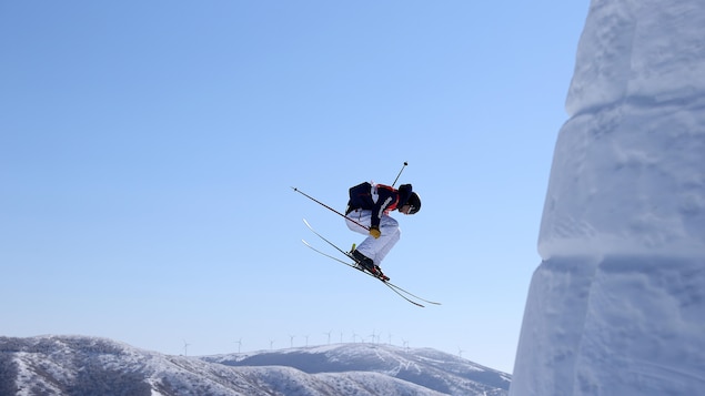 Un skieur acrobatique effectue un saut devant un château de neige. On voit des montagnes et des éoliennes loin derrière.