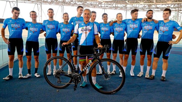 Sylvan Adams est à l'avant-plan d'un groupe de cyclistes. Ils sont tous vêtus en bleu. Adams a les mains sur un vélo.