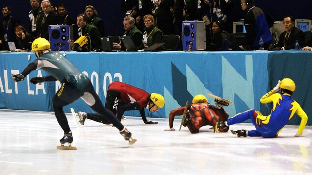 Un patineur de vitesse patine vers l'avant tandis que trois autres patineurs sont couchés sur la glace.