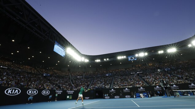 Un joueur au service dans un stade rempli, le soir.