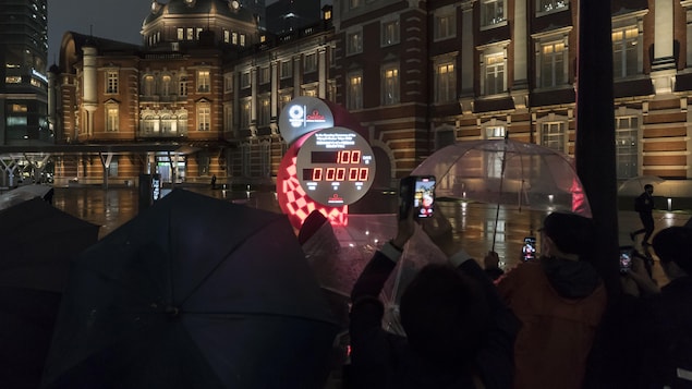 Une foule rassemblée sous des parapluies observent dans la pénombre le cadran indiquant le nombre de jours avant les Jeux olympiques de Tokyo.