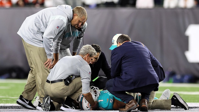 Allongé au sol, un joueur de football est soutenu au niveau du cou par un thérapeute sportif.