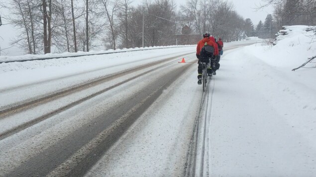 La route est enneigée. Des cyclistes sont sur le bord de la rue, en rang.