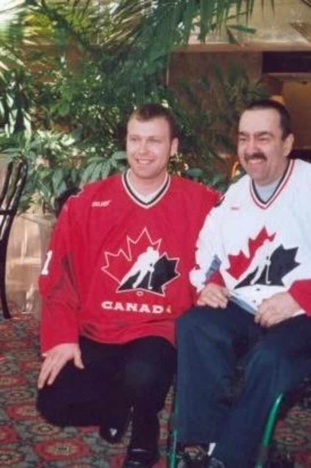 Martin Brodeur est accroupi à côté de Pierre Pichette, assis dans un fauteuil roulant. Les deux hommes portent un chandail de l'équipe canadienne de hockey.