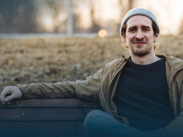 Un homme est assis sur un banc de parc et regarde la caméra en souriant légèrement.