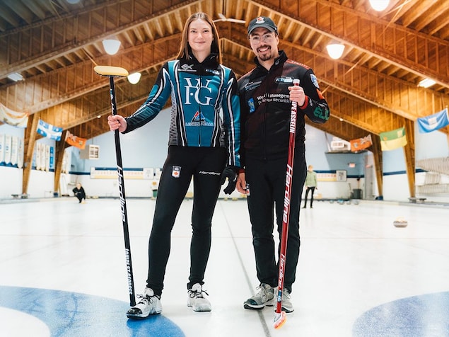 Deux joueurs de curling - un homme et une femme - sourient sur la glace, leur balais à la main.