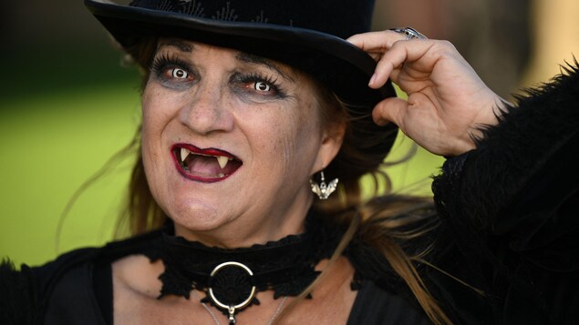 Une personne déguisée en vampire, avec un chapeau, des yeux d'une couleur très claire et des crocs.