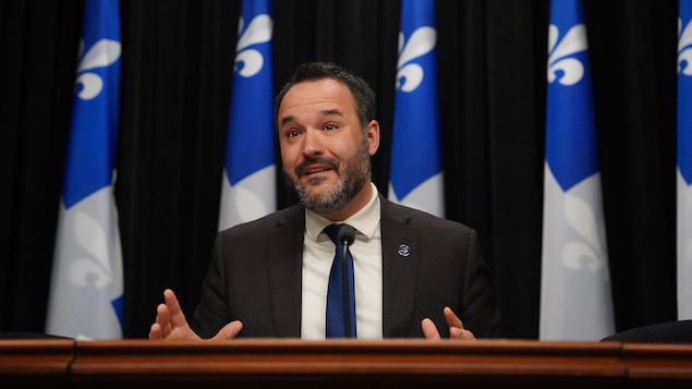 Le ministre s'exprime devant des drapeaux du Québec.