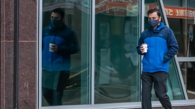 Un homme marche dans la rue, tenant un café dans la main, portant un masque. 