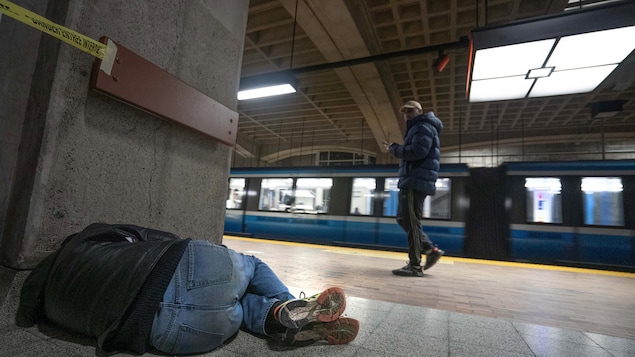 Un homme qui marche sur le quai du métro regarde un itinérant couché par terre, recroquevillé le long d'un mur.
