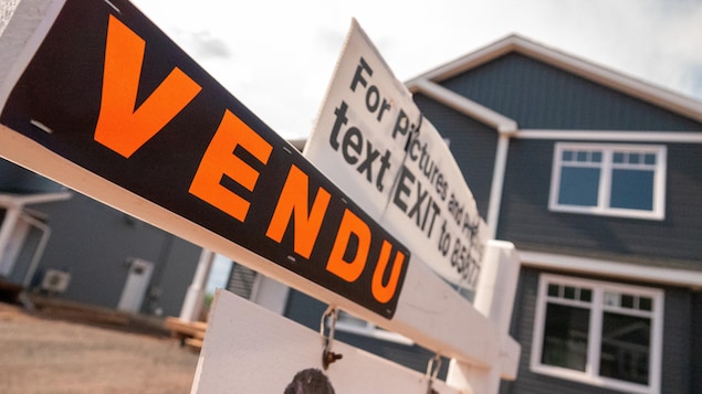 Affiche noire sur laquelle est écrit le mot « Vendu » en lettres orange devant une maison.