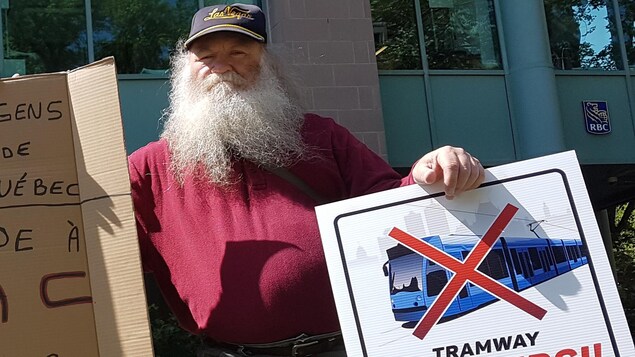 Un homme tient deux pancartes dont l'une représente un tramway barré.