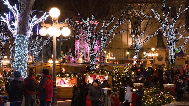 Des clients se tiennent dans le marché de Noël à la noirceur, entourés d'arbres illuminés de lumières de Noël.