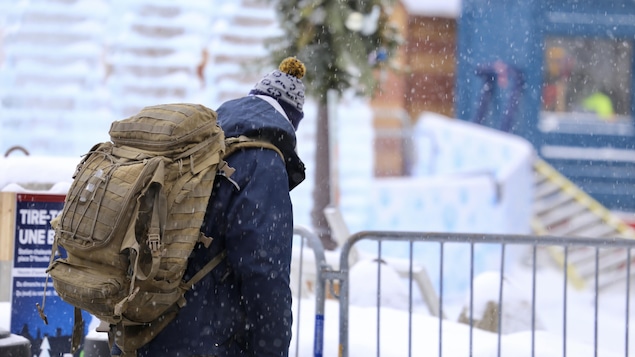 Une personne marche dans la rue sous la neige, de jour. Elle porte un sac à dos d'expédition.