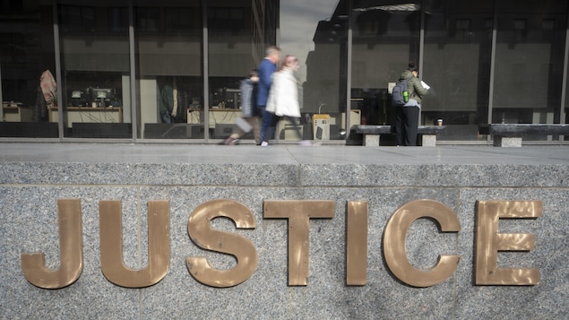 Un couple avec un enfant marche et une femme portant un sac à dos regarde une feuille devant le palais de justice de Montréal, avec à l'avant-plan le logo «Justice».