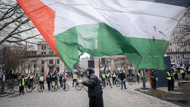 Une rangée de policiers est postée devant la grille de l'Université McGill pendant qu'un manifestant, le visage partiellement recouvert d'un keffieh, brandit un drapeau palestinien.