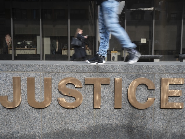 Une personne à l'avant-plan et une femme en arrière-plan marchent devant l'édifice du palais de justice de Montréal, au-dessus du logo «Justice».