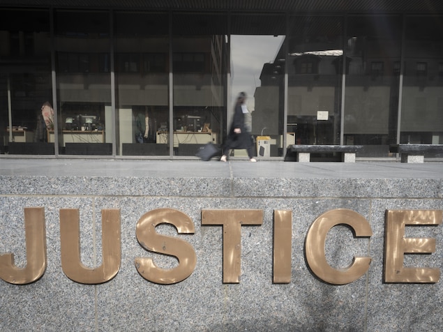 Une avocate en toge marche devant l'édifice du palais de justice de Montréal avec en avant-plan le logo «Justice».