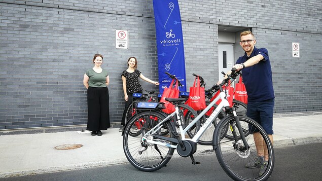 Un homme souriant tient un vélo électrique; deux femmes sont derrière lui à côté d'une affiche.